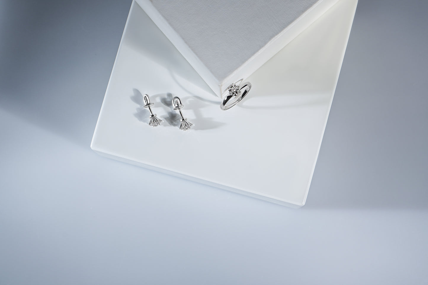 Сережки пусети та каблучка з діамантами у формі квітки. Зображені на білій підставці