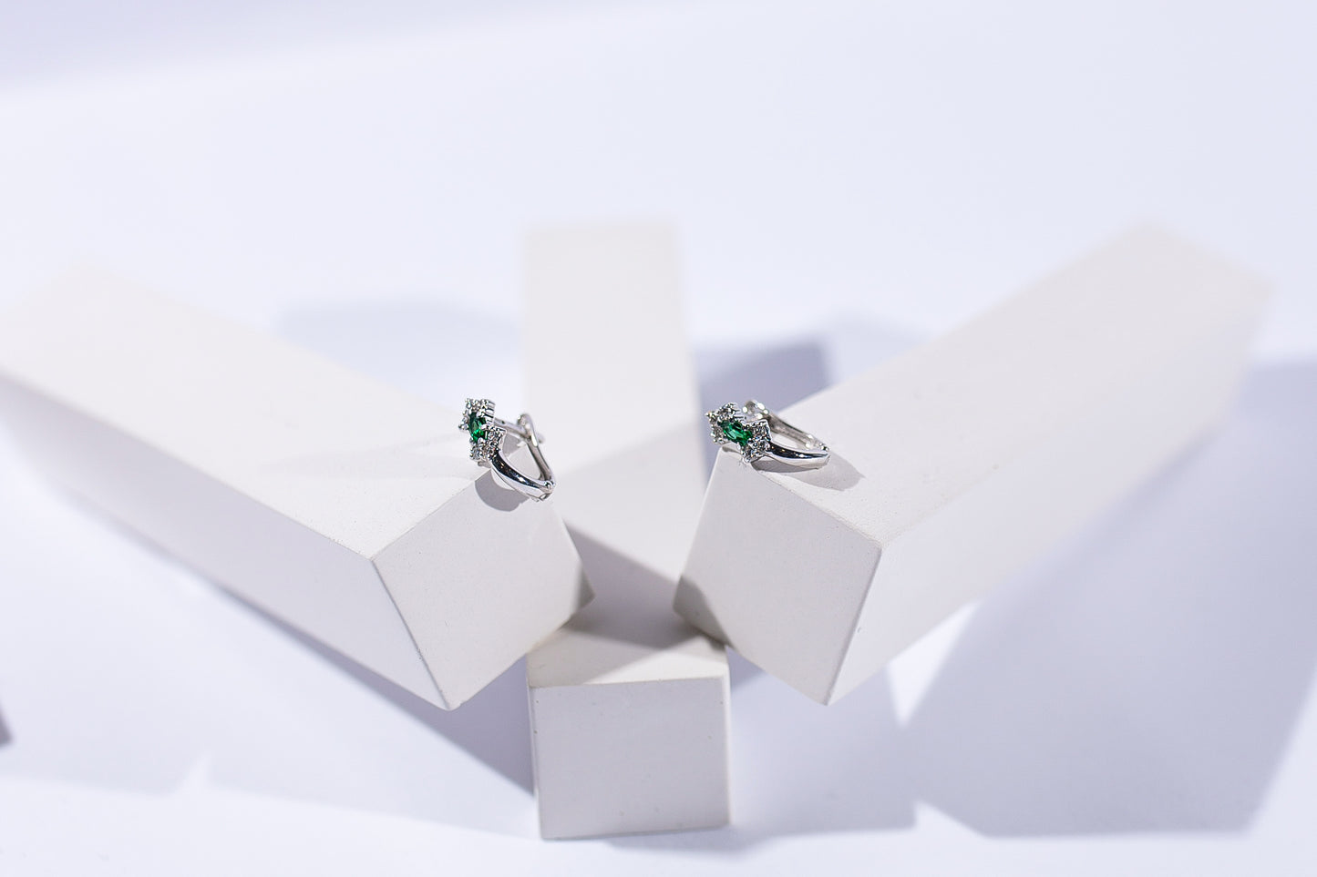 Дитячі сережки з білого золота з англійською застібкою зі смарагдом та діамантами. Зображені на білих прямокутних підставках