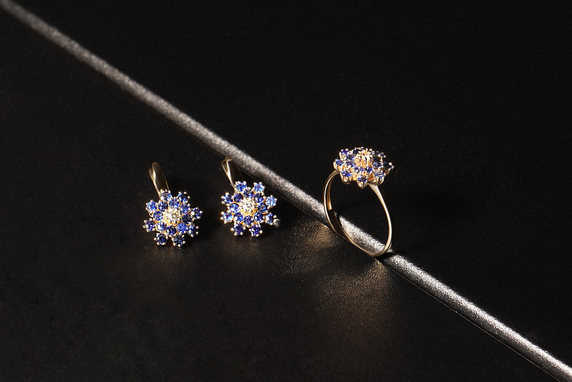 Набір сережки та каблучка з синіми сапфірами та діамантом по центру у формі квітки. Зображений на чорному фоні