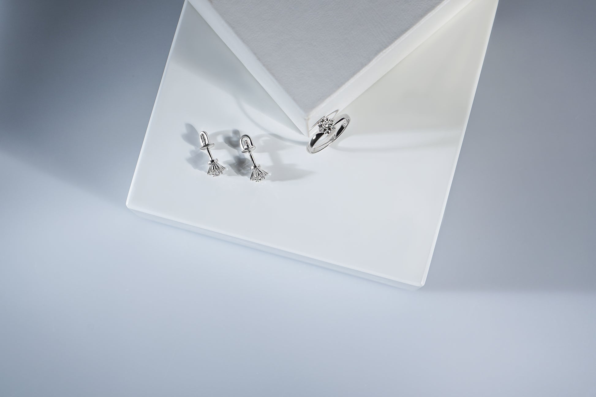Сережки пусети та каблучка з діамантами у формі квітки. Зображені на білій підставці