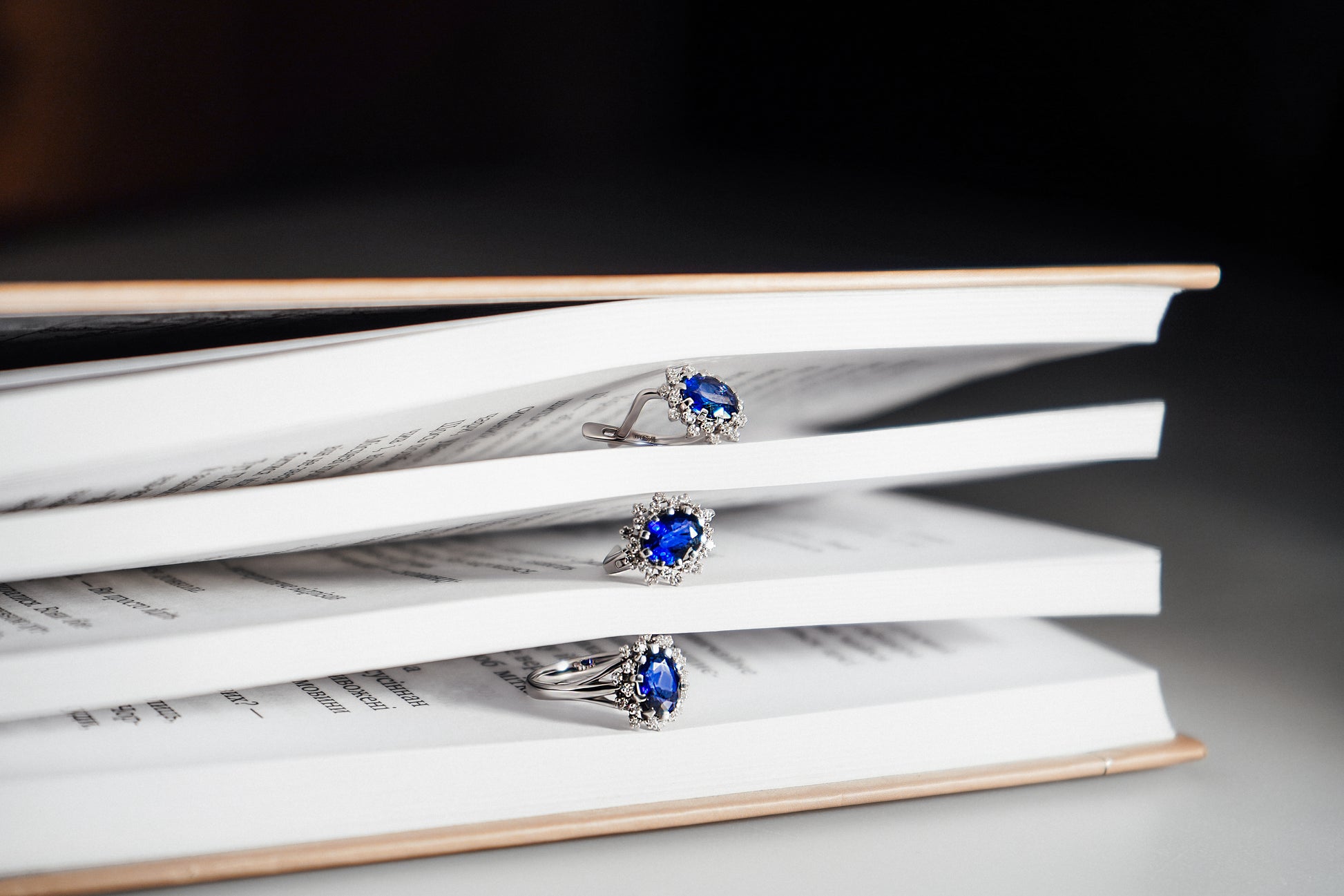 Набір каблучка та сережки з білого золота з великим синім сапфіром, навколо якого маленькі діаманти. Зображений між сторінками книги.